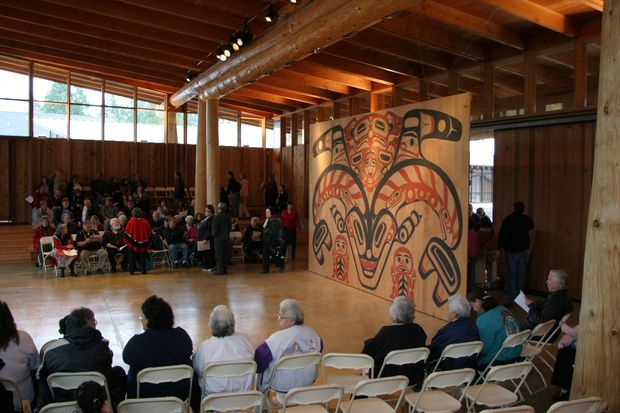 Large timber-framed gathering hall with exposed wood beams, columns, and ceiling structure. Rows of folding chairs face a large freestanding or partition-like Indigenous-inspired graphic artwork, with many people gathered for an event. Tall windows line the left side, bringing in daylight, while the polished wood floor and warm timber interior dominate the space.