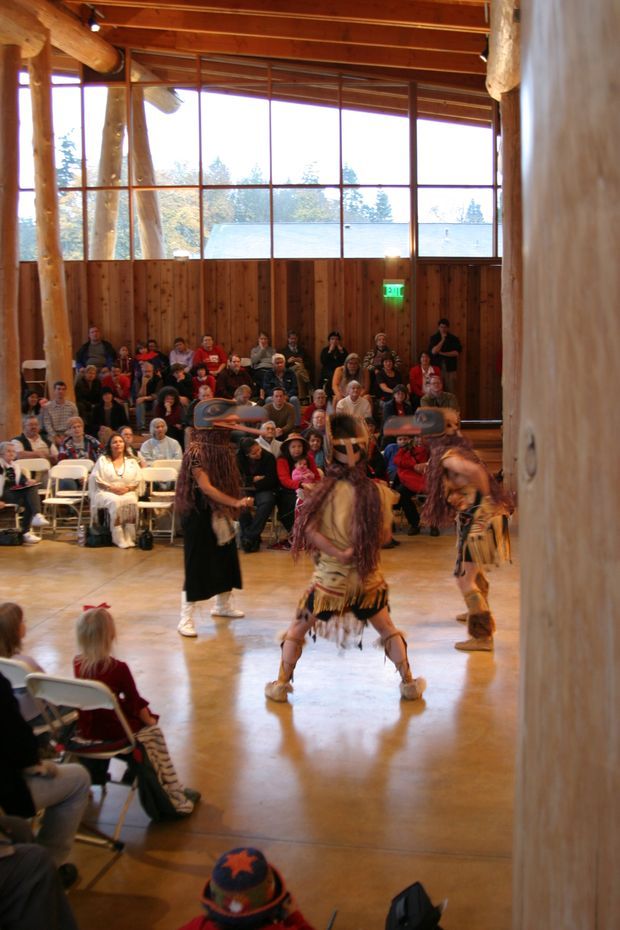A large timber-lined hall with exposed round-log posts and beams, warm wood ceiling, polished concrete floor, and extensive grid-framed glazing. An audience sits on folding chairs around performers in traditional dress, suggesting a community or cultural gathering space.