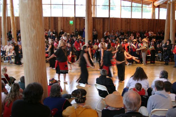 A large timber-lined hall with polished wood flooring, exposed round timber posts, high windows, and a crowd seated around the perimeter watching a cultural dance performance. Dancers and musicians occupy the central open floor, creating a community gathering atmosphere.