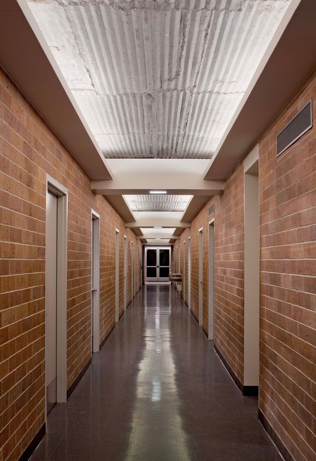 Long institutional hallway with exposed reddish brick walls, multiple closed doors, polished dark concrete flooring, and a repeated series of recessed ceiling panels containing corrugated concrete or metal inserts and linear lights. The corridor has strong symmetry, deep perspective, and an industrial, utilitarian character.