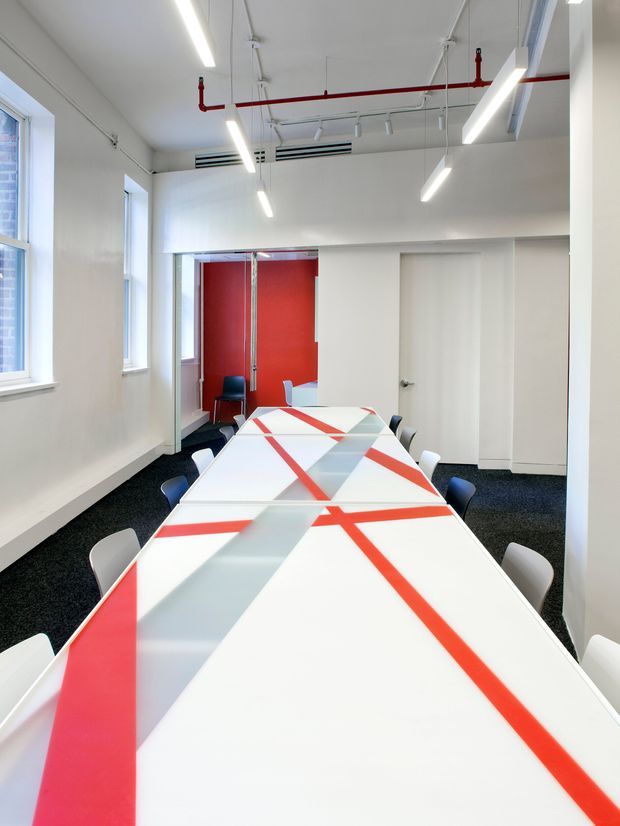 A bright contemporary commercial meeting room with a long white conference table surrounded by mixed white, grey, and dark blue chairs. The table features bold translucent panels and red linear accents. White walls, large windows, exposed ceiling services, red fire-sprinkler pipework, linear LED lighting, dark carpet, and a vivid red glazed partition create a clean modern office aesthetic.