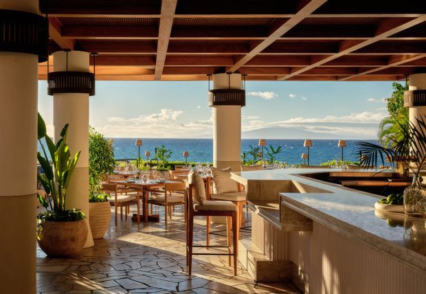 A spacious covered outdoor terrace overlooking the ocean, furnished with a dining table and chairs beside a substantial bar or outdoor kitchen counter. Large round columns support a timber-framed ceiling with exposed dark wood panels and integrated pendant lighting. Stone-patterned paving, tropical plants, warm timber furniture, and the expansive sea view create a refined resort-like setting.