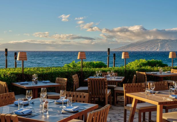 Outdoor waterfront dining terrace with multiple wooden tables and chairs set for meals, shaded by freestanding lamps and bordered by dense greenery. The terrace overlooks deep blue water toward a mountainous coastline under a partly cloudy sky.