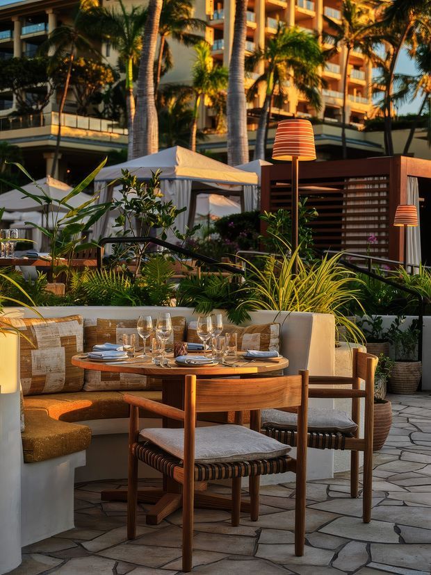 A warmly lit outdoor dining terrace at a resort or hospitality property, with a round timber dining table set with plates and wine glasses, upholstered timber chairs, a curved banquette, tropical planting, decorative outdoor lamps, cabana structures, and a multi-storey building in the background. The space has a refined tropical resort atmosphere and irregular stone paving.