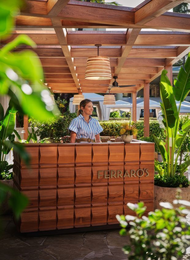 A tropical outdoor hospitality reception or bar counter beneath a timber pergola. The scene features a richly finished geometric timber front desk with the word “FERRARO’S,” warm pendant lights, a ceiling fan, lush plantings, and glimpses of outdoor seating and umbrellas beyond. The image has a warm, resort-like atmosphere with strong timber detailing and integrated greenery.