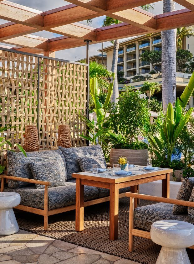 A bright tropical outdoor lounge and dining area beneath a timber pergola with translucent roof panels. The space features woven privacy screening, timber-framed upholstered lounge chairs and sofa, a small timber dining table, patterned outdoor rug, ceramic side stools, and abundant tropical planting. A multi-storey resort-style building is visible beyond the garden.