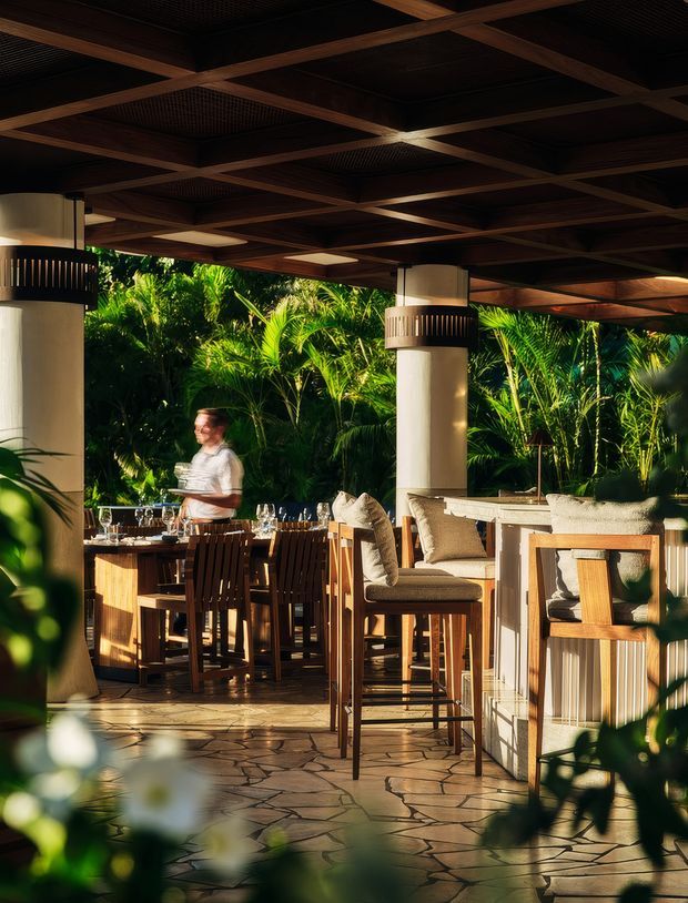 A warmly lit, open-air dining and lounge terrace surrounded by lush tropical planting. The space features a dark timber open-grid ceiling, substantial round columns with decorative dark banding, irregular natural stone paving, timber dining and bar-height furniture, upholstered seating, and a built-in outdoor counter or bar. The setting has a resort-like, tropical atmosphere with strong indoor-outdoor connection.