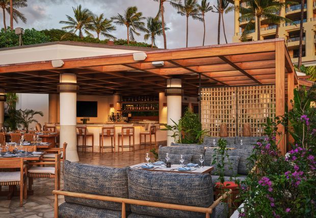 A tropical open-air restaurant or resort lounge with a timber pergola roof, warm ambient lighting, bar and high stools, dining tables, woven outdoor seating, and a decorative timber lattice screen. Palm trees, lush planting, and a multi-storey building are visible in the background.