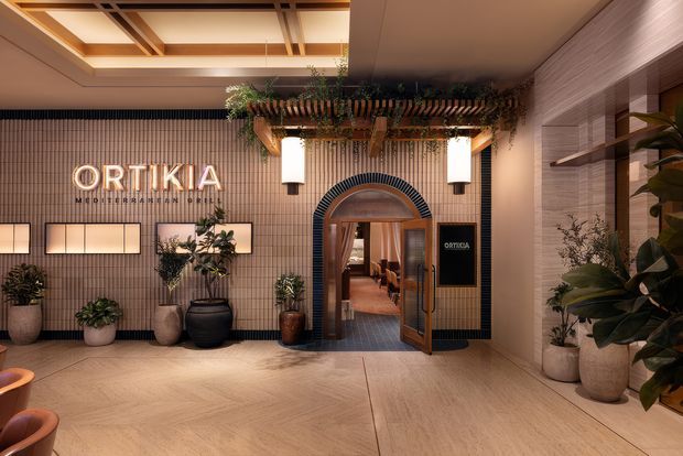 A refined hospitality entrance lobby for Ortikia, featuring a tiled feature wall with illuminated signage, a rounded arched doorway framed in dark ribbed tile, timber-and-glass entry doors, integrated wall lighting, planted pots, and a slatted overhead canopy with trailing greenery. The space uses warm timber-toned flooring, textured neutral walls, and layered architectural lighting.