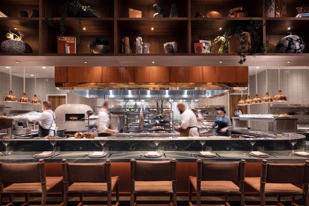 A warmly lit contemporary restaurant interior viewed from the dining counter, with chefs working in an open kitchen behind a long stone counter. The space features dark timber shelving and cabinetry, stainless-steel cooking equipment, tiled walls, pendant and recessed lighting, and rows of leather-upholstered counter stools. Decorative objects and plants are displayed on the upper shelving.