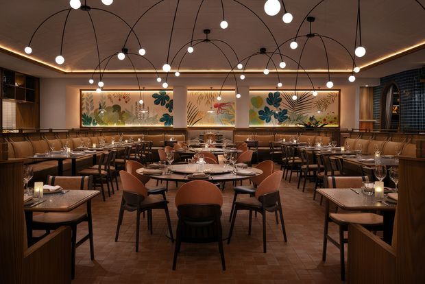 A richly styled restaurant dining room with numerous round and rectangular tables, upholstered dining chairs, dark timber wall panelling and cabinetry, and large colourful botanical artworks. The ceiling features a dramatic web of curved black pendant arms with glowing globe bulbs, while warm concealed lighting creates an intimate, sophisticated atmosphere. The floor appears to be laid with small terracotta or reddish-brown tiles in a patterned arrangement.