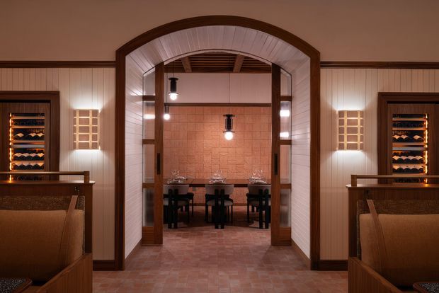 A warmly lit, symmetrical interior showing a wine-focused dining or tasting area viewed through a large arched opening. Two built-in wine display cabinets flank the foreground, with upholstered booth seating, wood trim, vertically panelled walls, and a central dining table set with glassware. The rear room features terracotta-toned tiled walls and flooring, dark wood details, and pendant lighting, creating an intimate hospitality-style atmosphere.