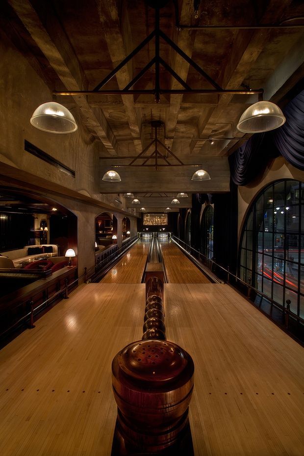 A dramatic interior view down a polished bowling alley in a dark, atmospheric entertainment venue. Multiple timber bowling lanes lead toward pins and a bar or lounge area, with warm wood surfaces, exposed timber ceiling beams, black metal trusses, industrial pendant lights, arched steel-framed glazing, and deep charcoal drapery. The space has a richly detailed, vintage-industrial character with low warm lighting.