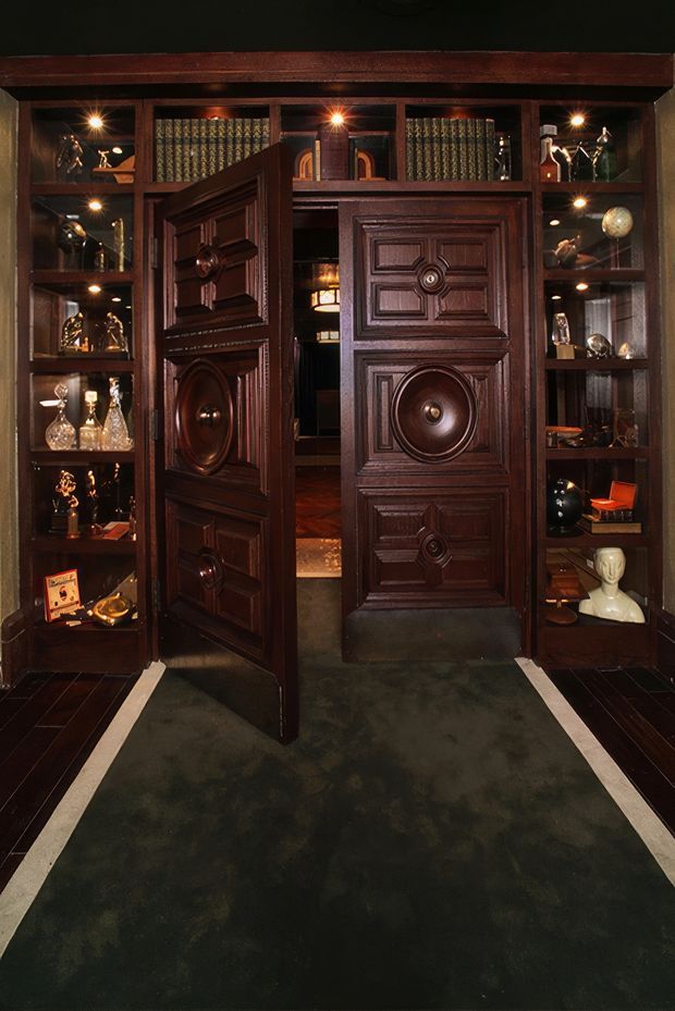 A dark, richly detailed interior entrance or display room centered on ornate dark timber double doors, one open and one closed. Built-in timber shelving on both sides contains decorative objects and glassware, with warm recessed spotlights illuminating the cabinetry. A dark carpeted floor with light border strips leads toward the doorway, and a glimpse of a warm wood-floored room is visible beyond. The overall atmosphere is dramatic, vintage, and highly ornamental.