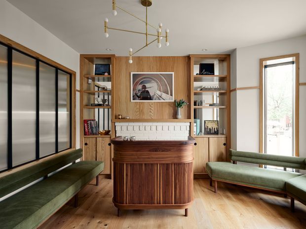 A warm, carefully detailed living room centred on a fluted timber bar or console beneath a framed artwork and white brick fireplace surround. Full-height timber built-in shelving and cabinetry flank the fireplace, displaying books and objects. Green upholstered built-in bench seating lines the dark-framed glazing on the left and continues along the right wall. The room has pale timber flooring, white walls and ceiling, and a sculptural brass-and-white pendant light. The overall composition blends mid-century forms with contemporary custom joinery.