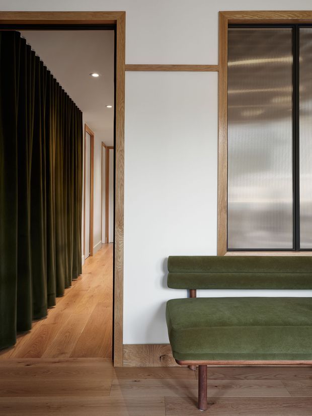 A refined interior hallway or entry area with warm oak framing, oak flooring and skirting, a deep olive-green upholstered built-in bench, and a black-framed textured translucent window. A dramatic dark olive curtain lines the corridor, creating a strong contrast with the white walls and timber detailing.