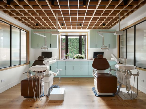 A bright, contemporary dental treatment room with two patient chairs and overhead dental equipment. The room features pale green built-in cabinetry, white walls, large black-framed glazed partitions, a central window, warm timber flooring, and a distinctive exposed timber coffered grid ceiling with integrated lighting.