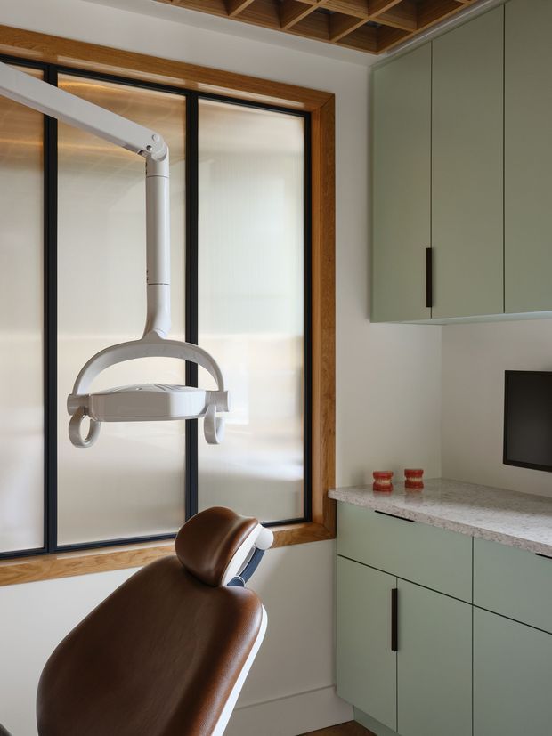 A compact, contemporary dental treatment room with a brown leather dental chair and overhead examination light. The room features large frosted-glass windows in dark frames with warm timber surrounds, pale sage-green built-in cabinetry, and a light speckled stone benchtop. A timber-lined coffered ceiling adds warmth and visual interest.
