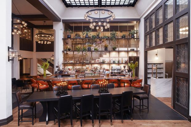 A sophisticated restaurant or cocktail bar interior with a long communal dining table in the foreground, a richly stocked bar with extensive open shelving at the centre, and multiple clusters of ornate pendant and chandelier lighting. The space features dark timber and black finishes, decorative metal-framed glazed partitions, patterned glass doors, warm timber flooring, and high ceilings with a large skylight or roof lantern. The overall atmosphere is moody, luxurious, and refined.