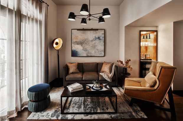 A sophisticated living room with a dark brown sofa, leather armchair, black-framed glass coffee table, patterned area rug, floor lamp, and a sculptural multi-light chandelier. Full-height sheer curtains bring soft daylight into the room, while a recessed display bar with glassware adds a refined focal point. Neutral taupe walls, dark timber flooring, and warm brown furnishings create an intimate, layered atmosphere.