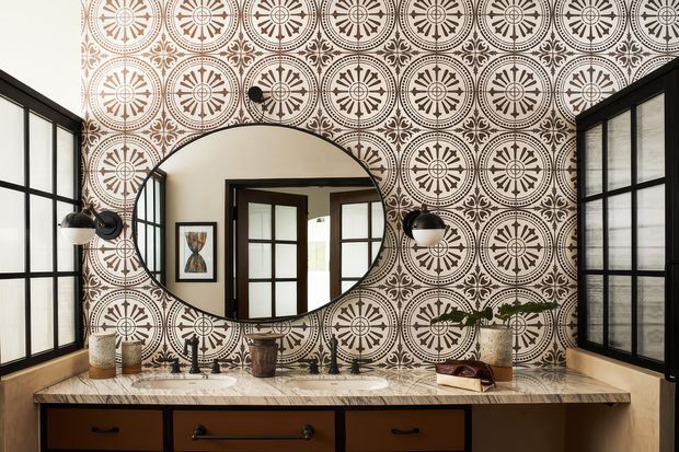 A richly patterned bathroom vanity with a long stone benchtop, twin basins, dark timber drawers, black-framed windows, wall sconces, and a large round mirror. The walls are covered in ornate black, cream, and terracotta geometric patterned tiles, creating a dramatic heritage-inspired interior.