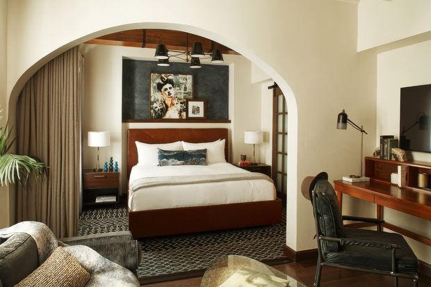 A warm, refined bedroom viewed through a large rounded archway. The room features a bed with white bedding and a dark wood bed base, patterned dark carpet, floor-length curtains, a dark feature wall with artwork, bedside tables and lamps, and exposed timber ceiling detailing. A timber desk with a leather office chair and wall-mounted television occupies the right side, while a sofa and coffee table are partially visible in the foreground.