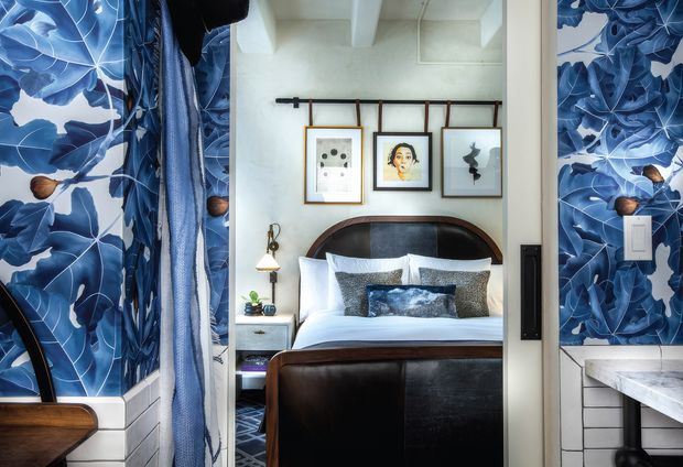 A compact bedroom viewed through a wide opening, featuring a dark brown leather upholstered bed with curved headboard and footboard, layered white and patterned bedding, and three framed artworks suspended from a simple wall-mounted rail. Bold blue botanical wallpaper and blue curtains frame the room. White painted walls, exposed ceiling beams, and a partial marble-topped tiled vanity or counter are visible at the sides. The overall look is richly patterned, eclectic, and refined.