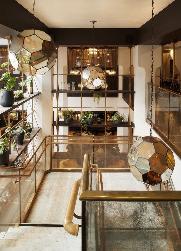 A dramatic multi-level interior atrium or stair landing with dark charcoal walls, cream ceiling panels, bronze-toned metal and glass balustrades, and extensive built-in shelving displaying potted plants and decorative objects. Large faceted geometric pendant lights hang at different levels, creating a warm, sculptural focal point. The space has an industrial-luxe character with exposed circulation zones, reflective glass, and warm metallic detailing.