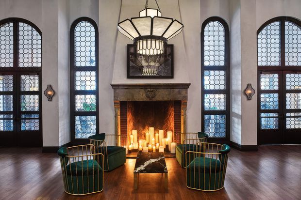 Grand, symmetrical interior room with a tall central masonry fireplace, dark timber mantel, glowing candles, and an ornate suspended geometric chandelier. Tall arched stained-glass windows and matching glazed doors line the walls, while deep green upholstered lounge chairs with brass-colored frames surround a low table on dark timber flooring. The atmosphere is dramatic, warm, and historic with a refined contemporary furnishing scheme.