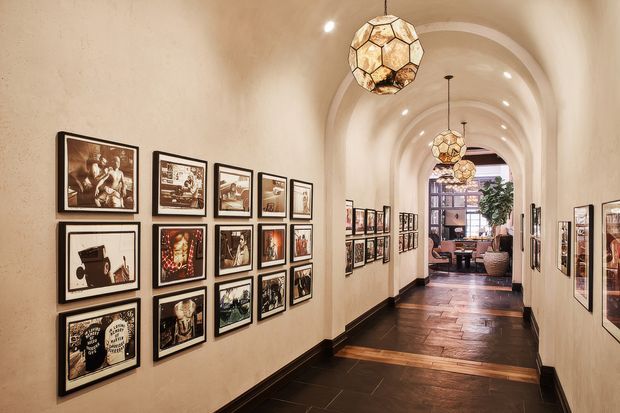 A long, elegant interior hallway or gallery passage with repeated rounded arch openings, cream textured walls, dark timber skirting, and dark wood flooring. The walls are densely arranged with framed artwork and photographs, while decorative geometric pendant lights hang along the vaulted corridor. A furnished sitting area and large window are visible at the far end.