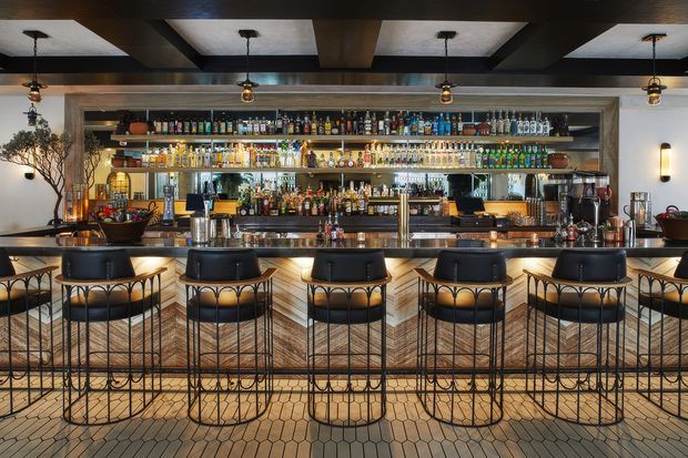 A sophisticated bar interior viewed straight on, with a long stone or concrete counter, a row of black upholstered bar stools with metal frames, and extensive backlit shelving displaying bottles and glassware. The space features dark exposed ceiling beams, warm pendant lighting, textured wall finishes, and patterned tiled flooring.