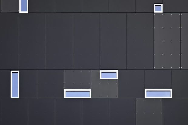 Abstract close-up of a dark charcoal modular wall or facade composed of rectangular panels with visible fasteners and several small, white-framed inset openings or illuminated blue-grey recesses. The image is tightly cropped and shows no identifiable room or broader building context.