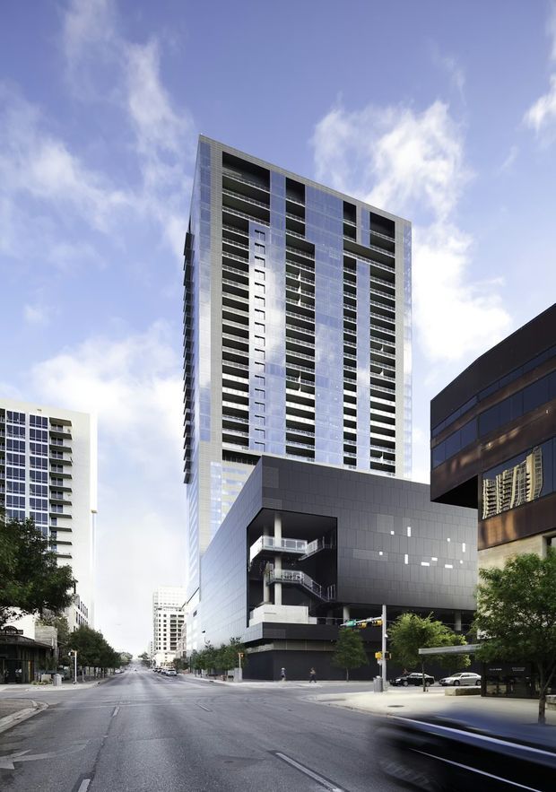 Street-level view of a tall contemporary urban high-rise complex, with a slender residential or mixed-use tower featuring repetitive balconies and a dark, angular podium. Additional multi-storey buildings line the broad city street, with trees, sidewalks, and moving traffic in the foreground.
