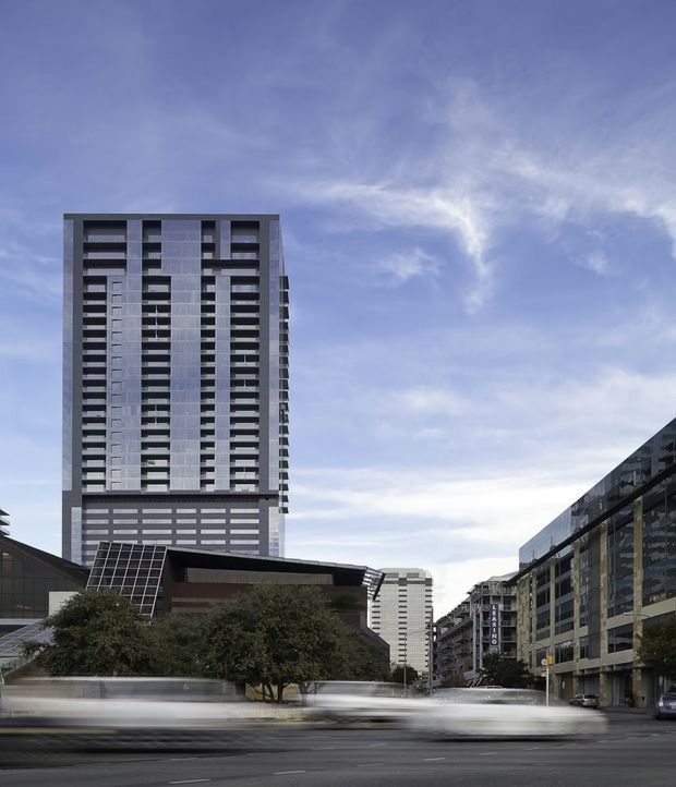 Urban architectural exterior photograph showing a tall modern high-rise tower with repetitive horizontal balcony or window bands, dark grey facade panels, and a lower podium. Additional multi-storey commercial or residential buildings line the street, with blurred traffic in the foreground and a partly cloudy sky.
