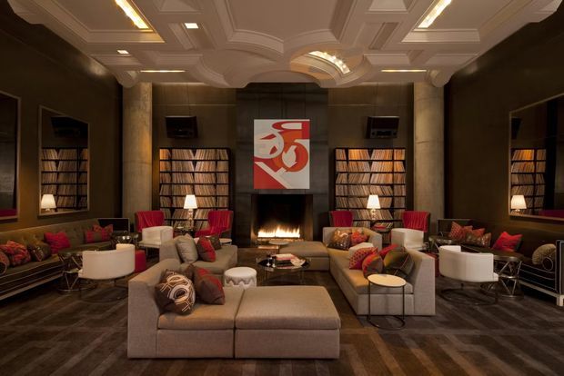 A sophisticated, warmly lit luxury living room or media lounge with a central fireplace, symmetrical built-in bookcases filled with books, wall-mounted televisions, dark timber wall finishes, and multiple seating groupings. The room features a large upholstered sectional, lounge chairs, ottomans, side tables, patterned cushions, and a highly ornate coffered and recessed ceiling with integrated lighting. A bold red-and-white artwork hangs above the fireplace.