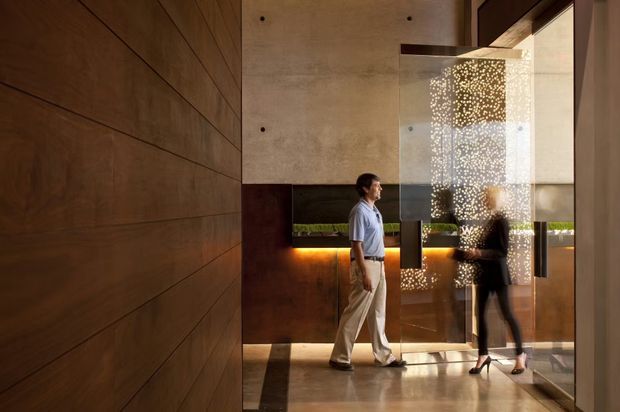 A sophisticated contemporary interior lobby or entry space with expansive dark timber wall panelling, exposed concrete, polished stone flooring, and warm concealed lighting. A decorative perforated or patterned glass screen divides the circulation area, with people walking through the space. The composition emphasizes layered materials, reflections, and a refined architectural fit-out.