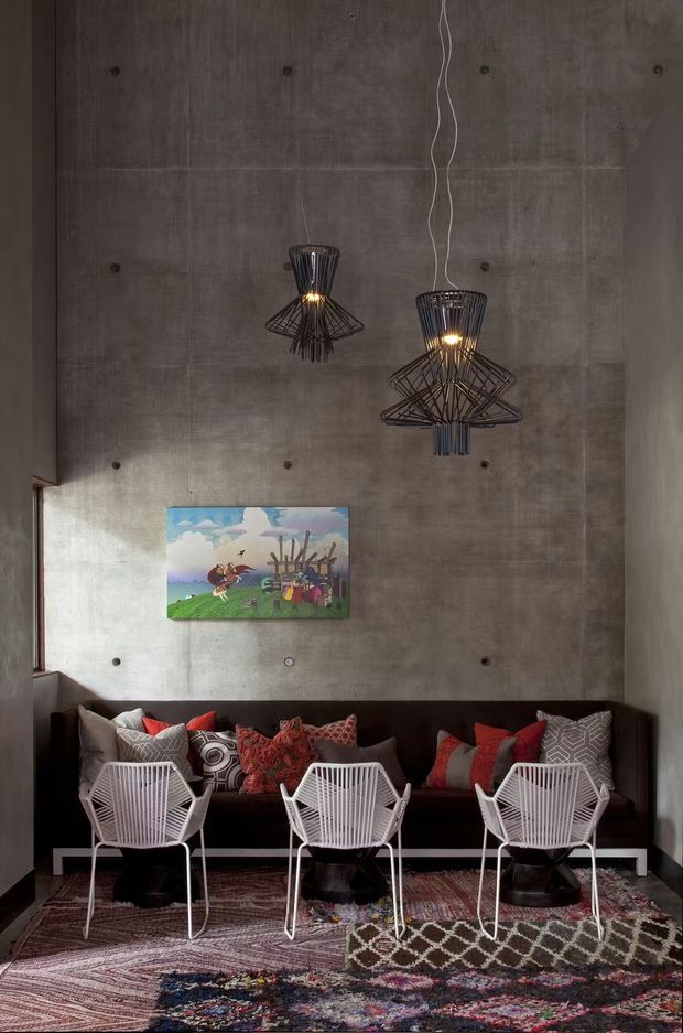 A dramatic contemporary lounge area with a raw poured-concrete feature wall, dark built-in bench seating, three white wire-frame lounge chairs, patterned cushions, and layered richly coloured rugs. Two sculptural black pendant lights hang from the high ceiling, while a colourful artwork provides a focal accent. The space has an industrial architectural shell softened by eclectic, graphic furnishings.