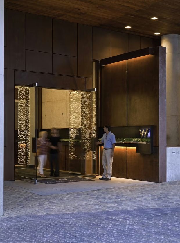 A sophisticated commercial entrance or hotel lobby frontage with dark bronze-toned panelled walls, a warm timber ceiling, recessed lighting, an illuminated reception or concierge counter, and a large pivoting glass door. Textured stone columns and patterned paving frame the entry, while people are visible inside and near the doorway.