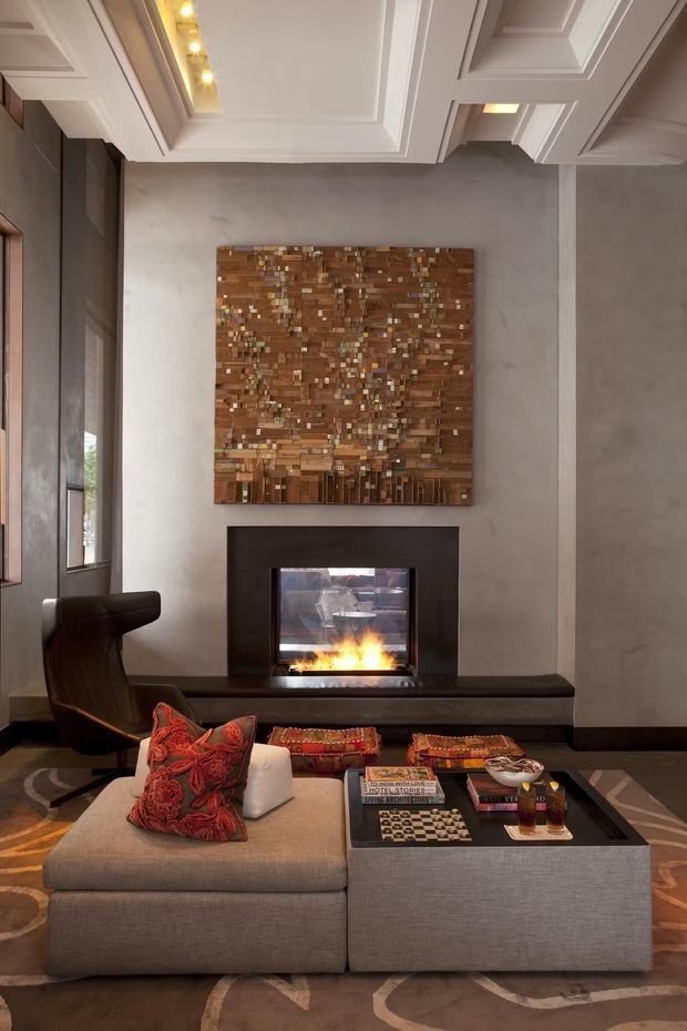 A refined living room centered on a contemporary fireplace with a dark stone or metal surround and a large textured wood mosaic artwork above it. The room features a low modular sectional sofa in warm grey upholstery, a dark square coffee table, a sculptural leather lounge chair, patterned cushions, and a coffered white ceiling with integrated lighting. Neutral plaster walls and dark timber accents create a sophisticated, warm atmosphere.