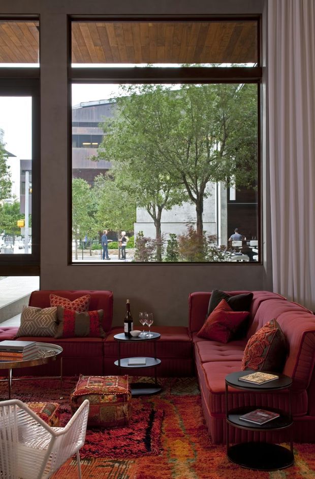 A richly layered living room with deep red upholstered sofas arranged around small black round coffee and side tables. Jewel-toned patterned cushions, a vibrant Persian-style rug, a woven ottoman, and a white woven lounge chair create an eclectic, warm interior. Large dark-framed windows overlook a leafy urban streetscape, while a timber-lined ceiling recess and full-height curtains add architectural and textural interest.