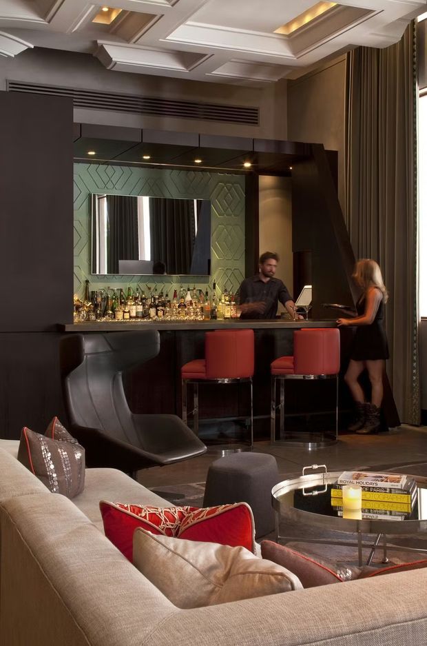 A sophisticated residential lounge with a built-in home bar at the rear. The dark bar cabinetry has a mirrored and patterned backsplash, illuminated bottle display, black countertop, and red upholstered bar stools. A large sectional sofa, sculptural lounge chair, and round coffee table occupy the foreground. The room features a highly detailed coffered ceiling with recessed lighting, dark curtains, and a dramatic palette of charcoal, taupe, red, and metallic accents.
