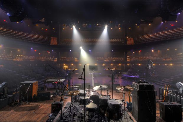 A large indoor concert or performance venue viewed from the stage, with a drum kit, microphones, keyboards, amplifiers, speakers, guitars, and other musical equipment in the foreground. The auditorium has extensive tiered seating, balconies, theatrical spotlights, and a dark atmospheric stage environment.