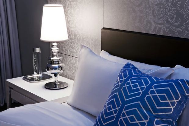 Close-up of a styled bedroom corner featuring a bed with white linens, a blue geometric-patterned accent cushion, a dark upholstered or timber-look headboard, patterned wallpaper, and a white bedside table with a chrome table lamp and accessories. Dark curtains frame the left side of the image.