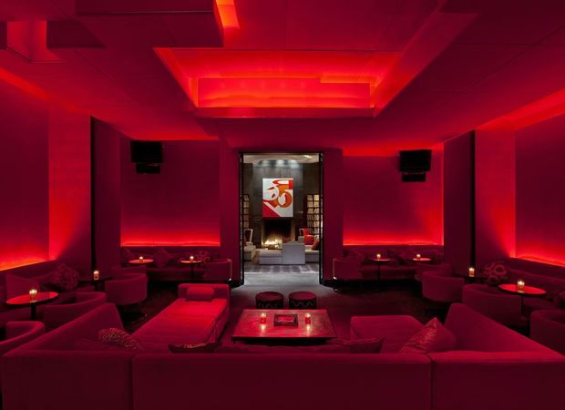 A dark, atmospheric lounge or entertainment room illuminated almost entirely in deep red. Low modular sofas and lounge seating surround small tables with candles, while wall-mounted speakers and recessed ceiling lighting reinforce a cinematic hospitality or home-theatre ambience. A doorway at the rear opens to another furnished space with a prominent artwork or sign.