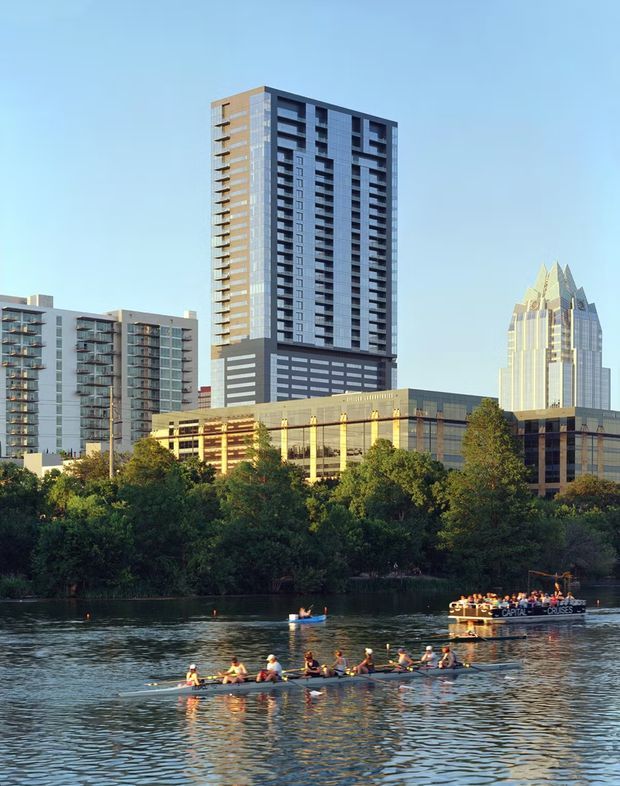 A daytime exterior view of a modern urban high-rise development beside a river. Several tall residential and commercial buildings with repetitive balconies, glazed façades, and strong vertical forms rise behind a tree-lined waterfront. Rowers and a sightseeing boat are visible on the water in the foreground.