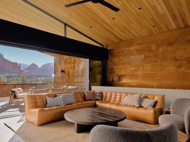 A warm, spacious contemporary living room opening onto an outdoor terrace, with a dramatic mountain landscape visible through expansive glazing. The room features a timber-lined ceiling with recessed downlights and ceiling fan, black-framed glazing and structural trim, a large rust-toned textured feature wall, tan leather sectional seating, sculptural upholstered armchairs, a round dark wood coffee table, and a large neutral rug. The space has a strong indoor–outdoor connection and a refined lodge-like atmosphere.
