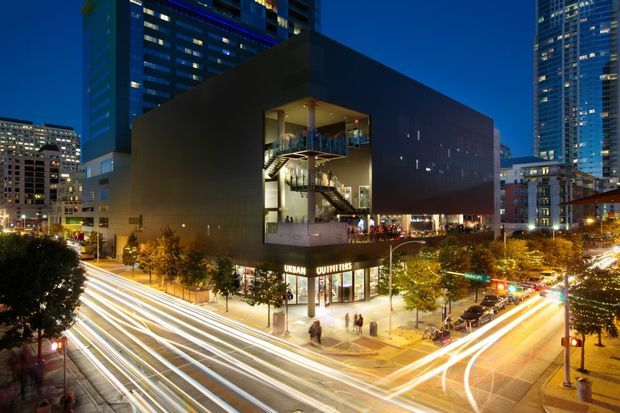 Night-time urban exterior photograph of a large contemporary commercial building at a busy city intersection. The dark, box-like facade features a dramatic multi-storey glazed opening revealing interior stairs and circulation spaces, with retail frontage at street level and illuminated high-rise buildings surrounding it. Long-exposure vehicle light trails emphasize the dense metropolitan setting.