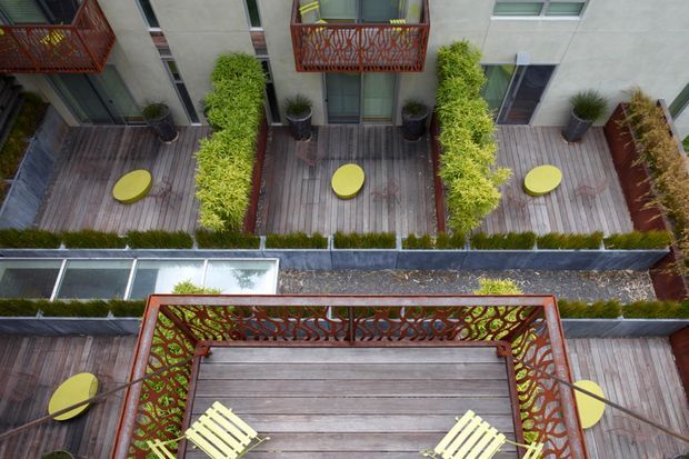 Aerial view of a multi-level urban residential courtyard or terrace complex. Several timber-decked outdoor terraces are arranged around planted garden beds, with bright yellow circular side tables and matching outdoor seating. Red-orange decorative metal balcony balustrades with organic patterned cut-outs contrast against pale concrete walls, dark decking, and abundant green screening plants.
