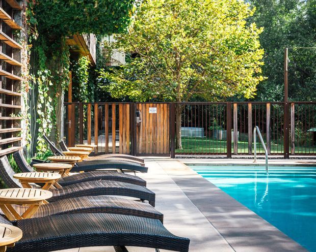 Outdoor poolside area with a row of woven chaise lounges and small timber side tables beside a bright rectangular swimming pool. Timber and dark metal fencing, dense greenery, climbing vines, and a pool access gate create a private resort-like setting. A slatted timber screen and warm timber structure are visible along the left side.