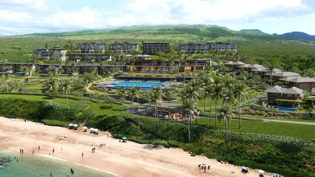 Aerial view of a large tropical beachfront resort with multiple low-rise buildings, landscaped gardens, swimming pools, palm trees, pathways, and a sandy beach beside the ocean. Green hills rise in the background under a partly cloudy sky.
